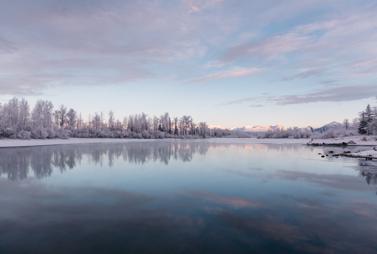 A picture of a lake in winter, with a forest in the back ground, at dusk.